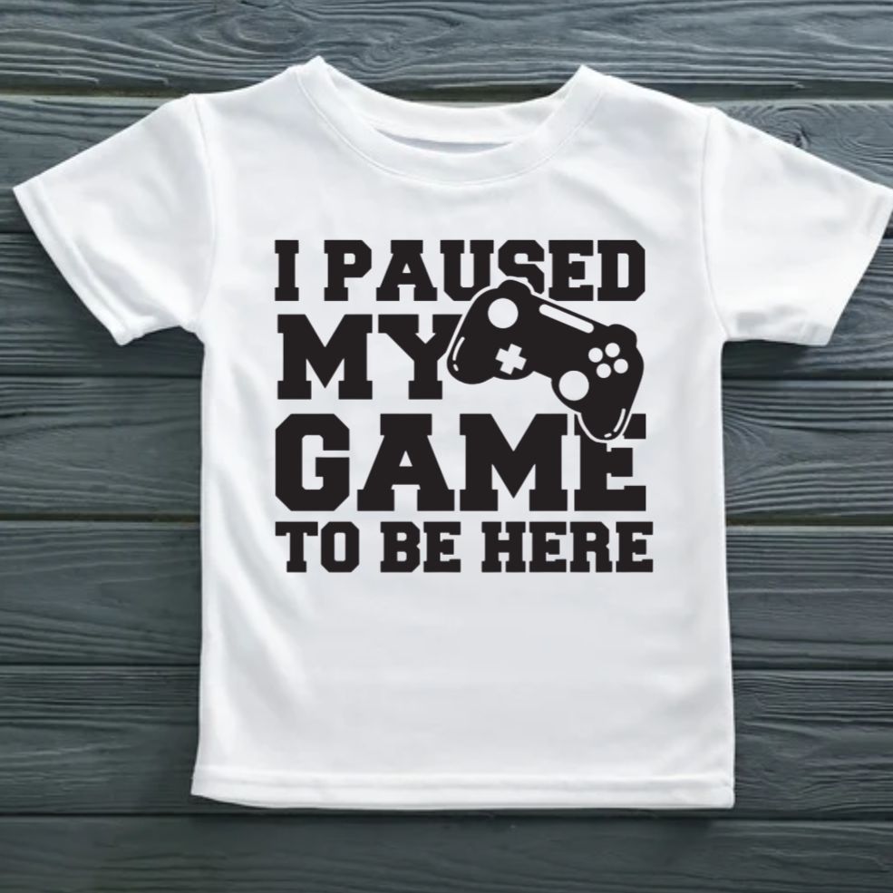 White t-shirt with black text and gaming controller graphic on a wooden surface