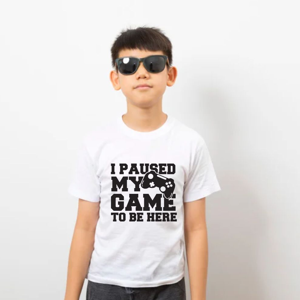 Child wearing a white t-shirt with 'I Paused My Game To Be Here' text and graphic on a plain background